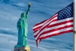 Hochauflösende Aufnahme der Freiheitsstatue mit US-Flagge im Wind, symbolisiert amerikanische Wirtsc.