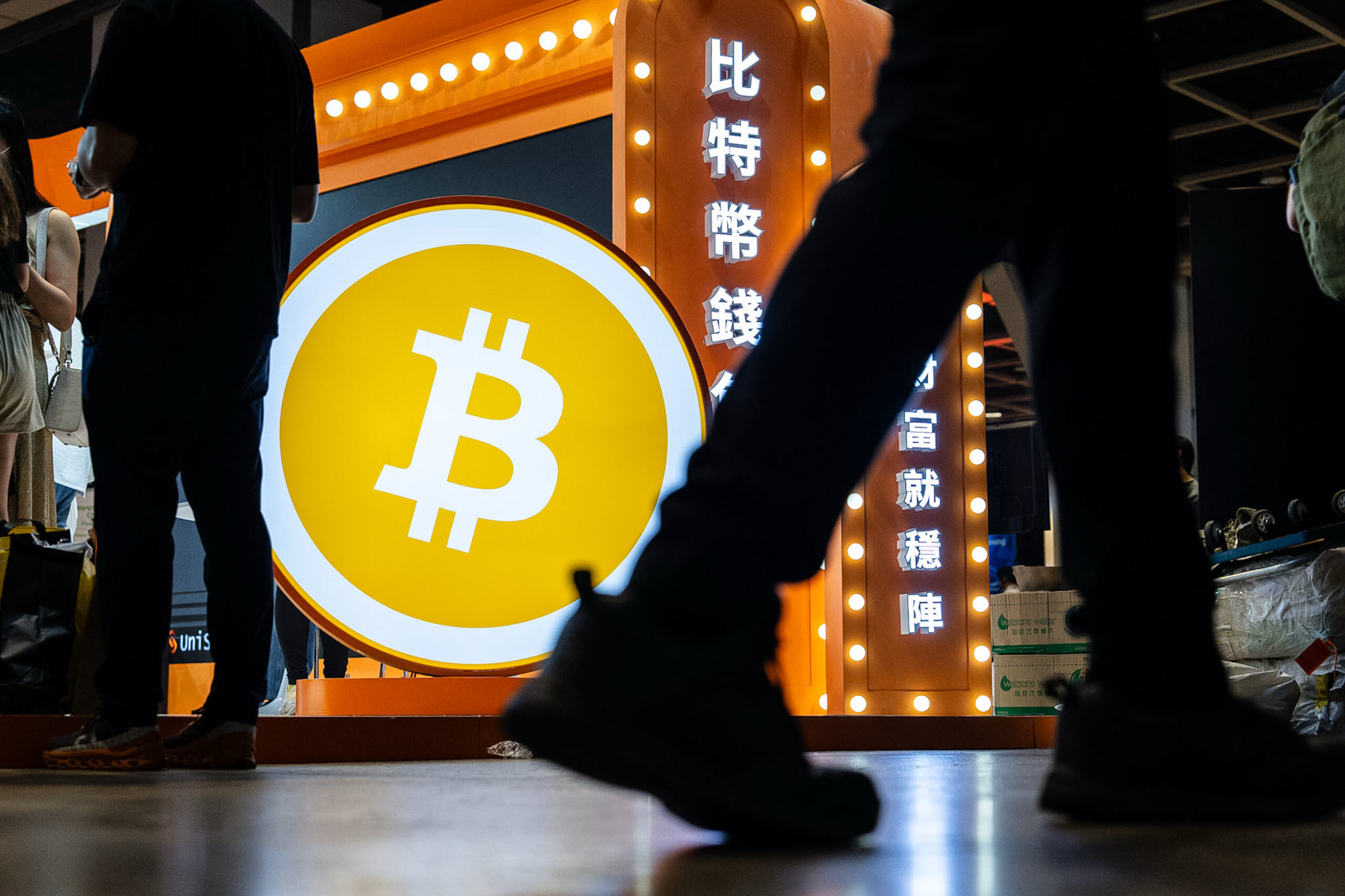 Bitcoin-Symbol auf einer Börsenmesse, Menschen im Hintergrund, Fokus auf Kryptowährung.