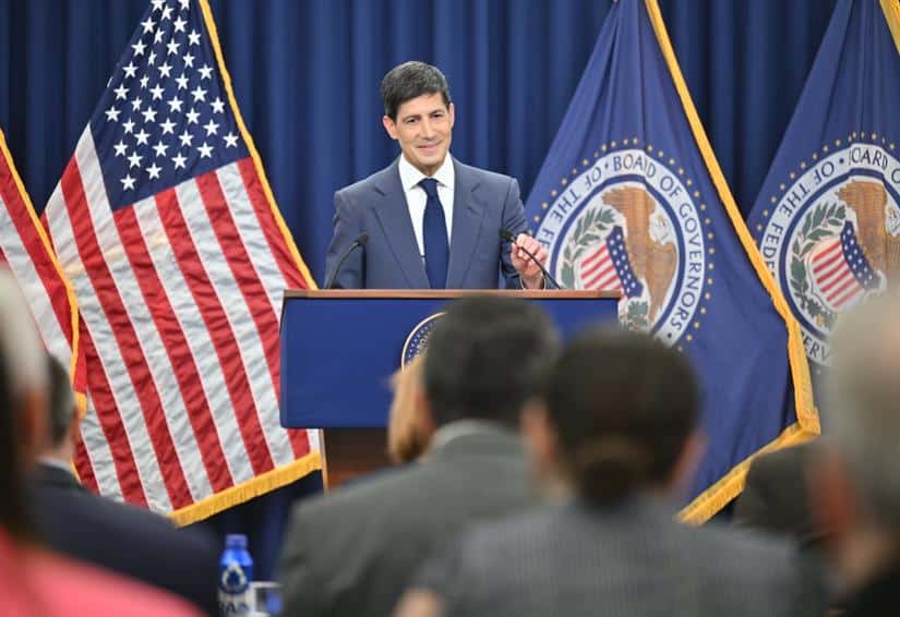 Fed-Chef Warsh bei Pressekonferenz vor US-Flagge und Fed-Flagge.